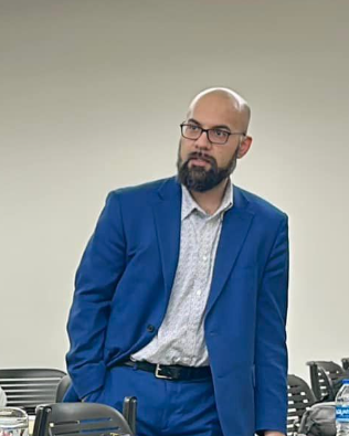 A man in a blue suit stands, looking thoughtfully in a meeting room.